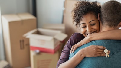 Junges Paar umarmt sich mit Schlüssel in der Hand. Im Hintergrund Umzugskartons. 