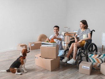 Ein Mann und eine Frau im Rollstuhl essen eine Pizza in einem leeren Raum mit Umzugskartons, ein Hund schaut ihnen dabei zu.