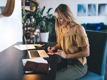 Frau sitzt am offenen Laptop und schreibt dazu einen Post-it