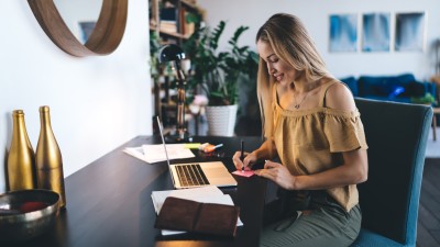 Frau sitzt am offenen Laptop und schreibt dazu einen Post-it