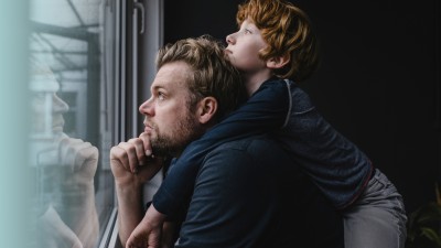 Mann sitzt nachdenklich vor einem Fenster und schaut aus diesem heraus. Ein Kind liegt auf seinen Schultern und schaut besorgt aus dem Fenster