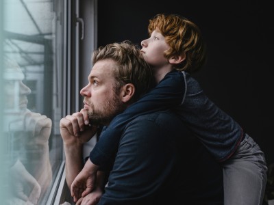 Mann sitzt nachdenklich vor einem Fenster und schaut aus diesem heraus. Ein Kind liegt auf seinen Schultern und schaut besorgt aus dem Fenster