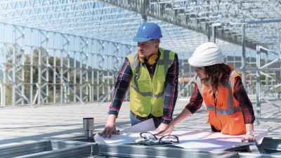 Ein Mann und eine Frau mit Schutzhelm und Warnwesten mit Plänen auf einer Baustelle