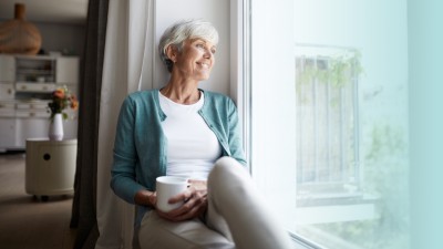 Ältere Dame sitzt lächelnd auf einer Fensterbank mit einer Tasse und schaut aus dem Fenster