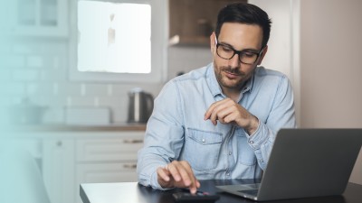 Mann sitzt vor einem Laptop und berechnet etwas auf einem Taschenrechner