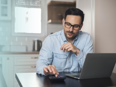 Mann sitzt vor einem Laptop und berechnet etwas auf einem Taschenrechner