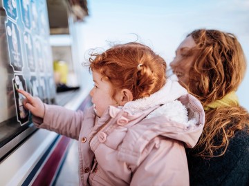 Kleines Kind in einer hellrosa Winterjacke zeigt auf die bebilderte Speisekarte eines Eiswagens an einem Verkaufsfenster, w&auml;hrend eine erwachsene Person danebensteht und ebenfalls auf das Angebot blickt.