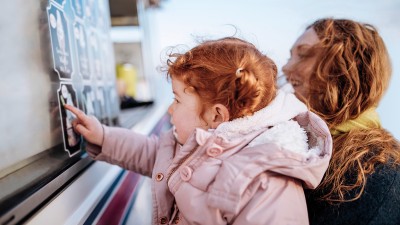 Kleines Kind in einer hellrosa Winterjacke zeigt auf die bebilderte Speisekarte eines Eiswagens an einem Verkaufsfenster, w&auml;hrend eine erwachsene Person danebensteht und ebenfalls auf das Angebot blickt.