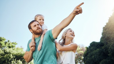 Frühstart-Rente für Kinder 2026: Ein Mädchen hängt am Rücken ihres Vaters, die Mutter steht daneben und stützt es