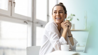 Junge Frau sitzt lächelnd vor einem Laptop mit einem Kaffee