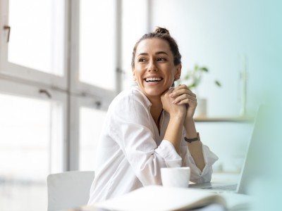 Junge Frau sitzt lächelnd vor einem Laptop mit einem Kaffee