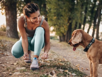 Gesundheit: Junge Frau kniet und bindet ihre Jogger-Schuhe und schaut dabei einen Hund an.