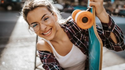 Zahn- & Brillenversicherung: Junge Frau lächelt und hält ein Skateboard fest