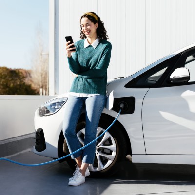 Elektroauto- & Hybrid-Versicherung: Junge Frau mit Ihrem Fahrzeug an der E-Ladestation