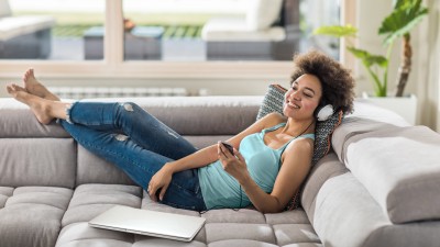 Hausratversicherung: Frau liegt auf Sofa mit Handy in der Hand und Kopfhörer auf dem Kopf